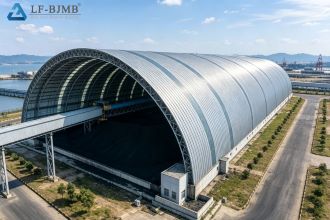 Aerial view of a completed large-span arched steel space frame coal storage shed, with belt conveyor gallery feeding the yard and coal pile visible inside the open end