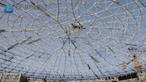 Space frame dome roof under construction, viewed from inside with crane visible in the distance.