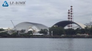 Two arched bolted ball-joint space frame coal storage sheds at a coastal power plant, one with cladding installed and one still as exposed steel grid